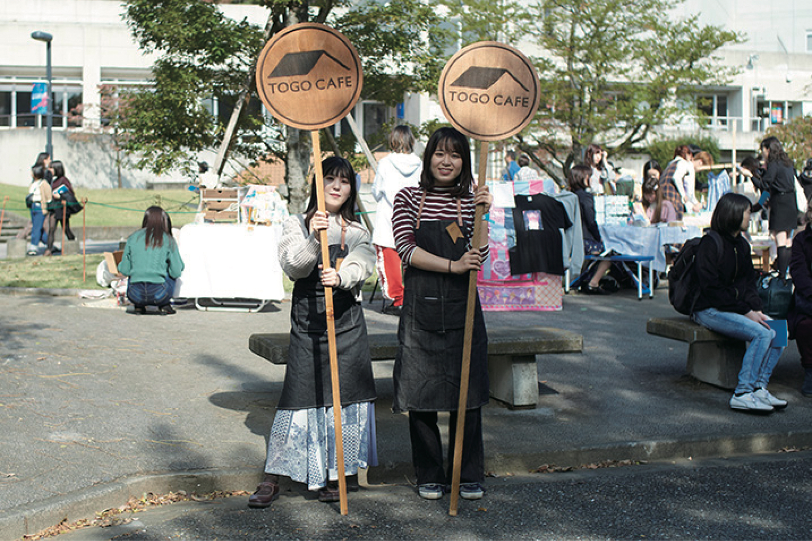 屋外イベントでカフェの看板を持つ女性二人が前に立っている様子。背景には人々が集まり、様々な物が売られている。