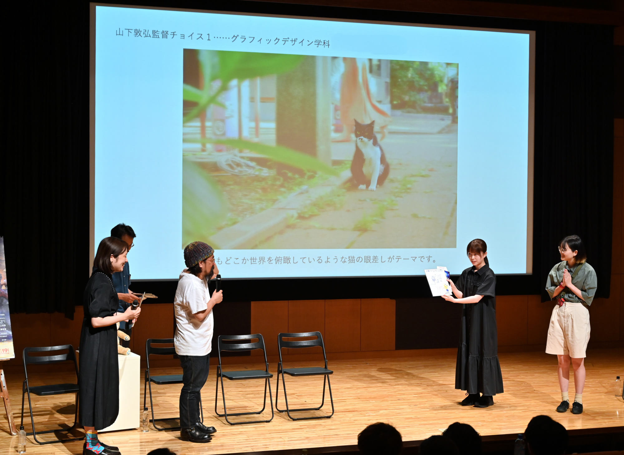 舞台上の大型スクリーンに久野監督と山下監督のお気に入り作品が投影され、前で学生が作品講評を受け記念のイラスト色紙を受け取っている。