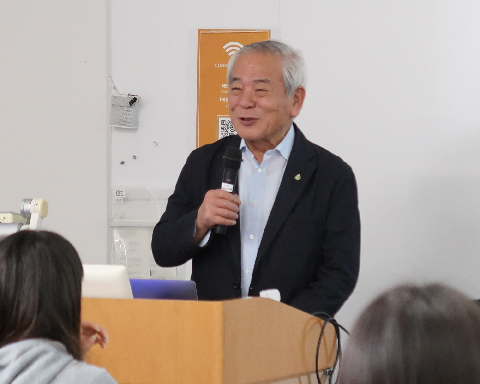 画面中央右手にマイクを持ち、笑顔で聴講する学生に語りかける青柳正規理事長。